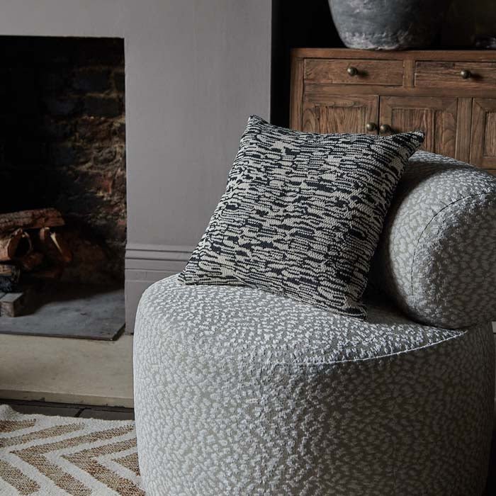 Woven fabric cushion cover in natural and black tones with an abstract pattern, sitting on a modern accent chair.