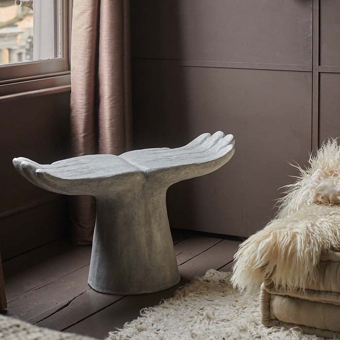 The Aimee Abstract Stool, a textured white resin stool in the shape of two open hands in a soft pink hued room by a white rug.