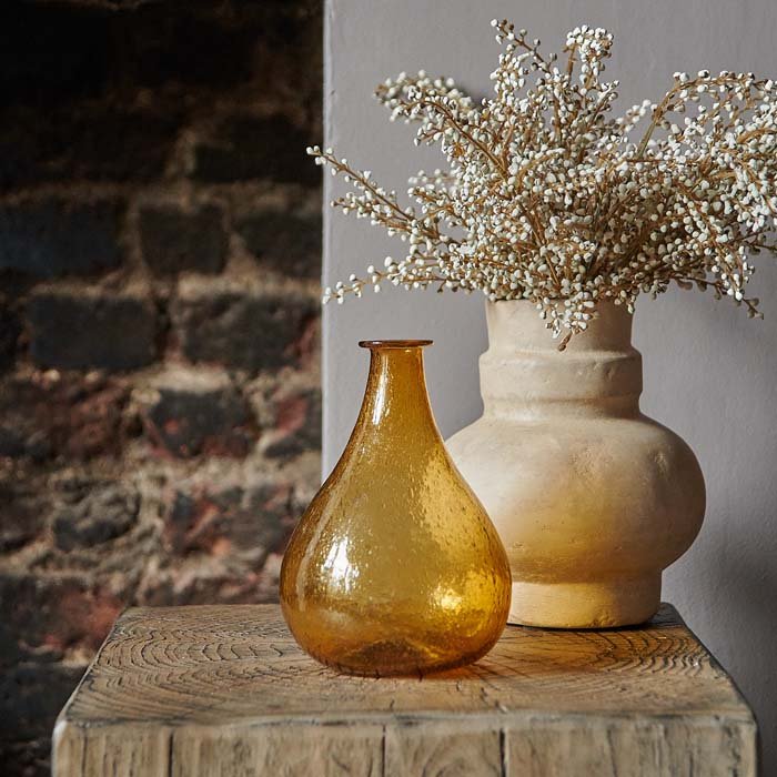 Styled image of a teardrop shaped amber glass vase next to a pale ceramic vase with a faux bouquet of flowers.