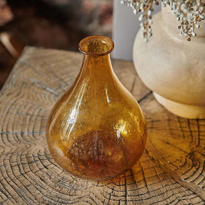 Amber glass bottle vase on wooden side table in an organic teardrop shape.