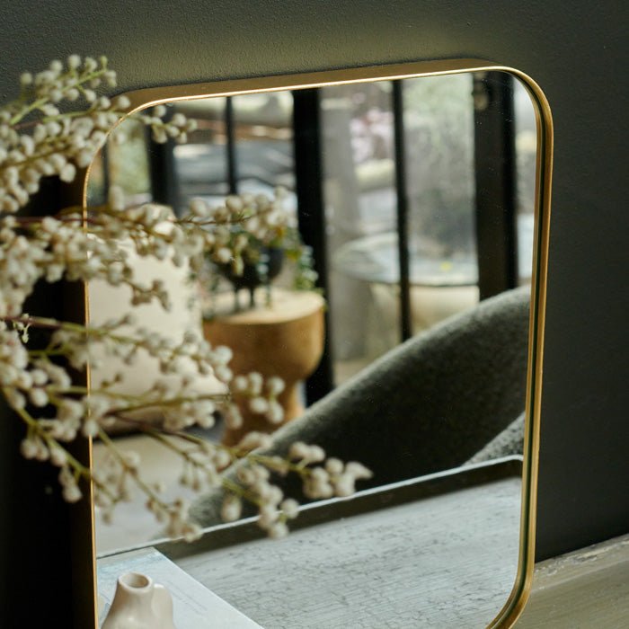 Gold framed square mirror with a curved edge leant against a green wall