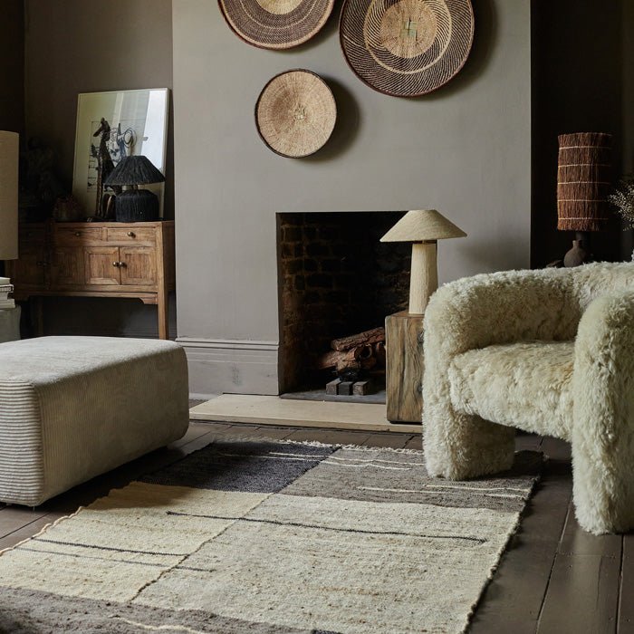 Woven rug with muted cream grey and navy linear block patterns next to a faux fur chair in front of a fireplace