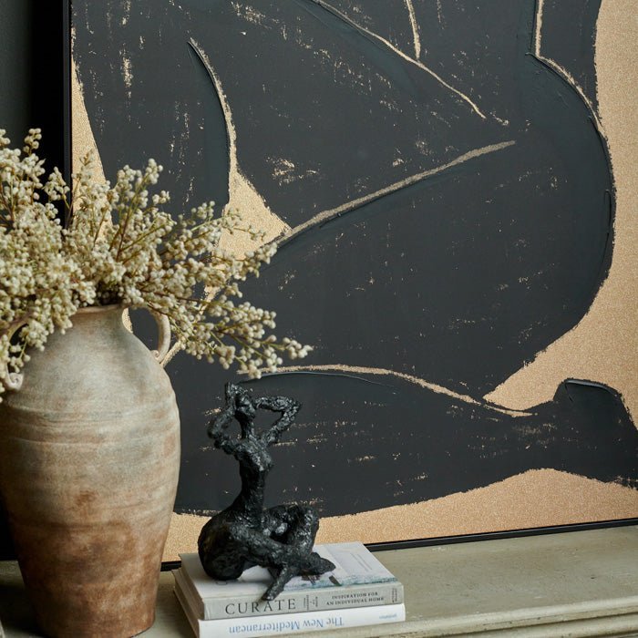 The Namora Canvas Print, a black and gold figurative canvas resting on a console behind a tall terracotta vase.