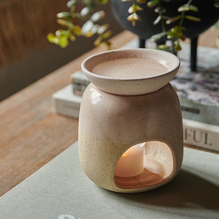 The Thea Oil Burner, a cream coloured ceramic oil burner with a lit candle inside, next to a book and a faux plant.