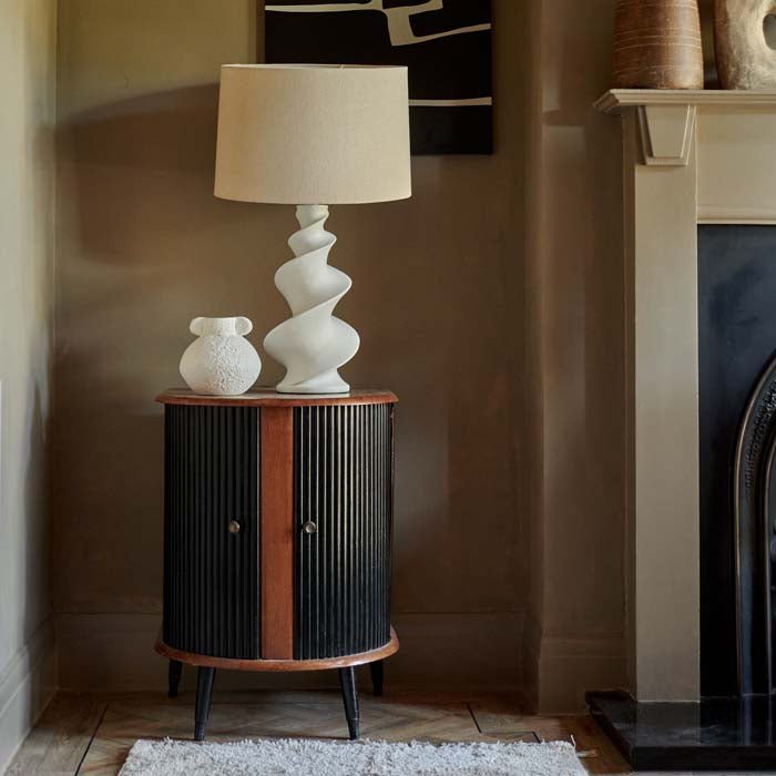 The Reo Table Lamp, a white lamp with a spiral white base and a cream drum shade, sat on a wooden side cabinet.