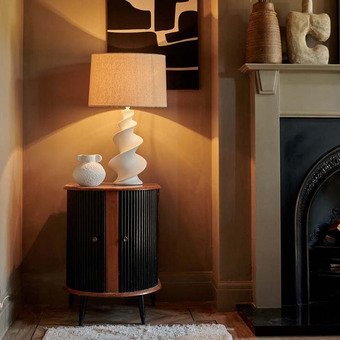 Reo Table Lamp, a white lamp with a spiral white base and a cream drum shade, warmly lit and sat on a wooden side cabinet.