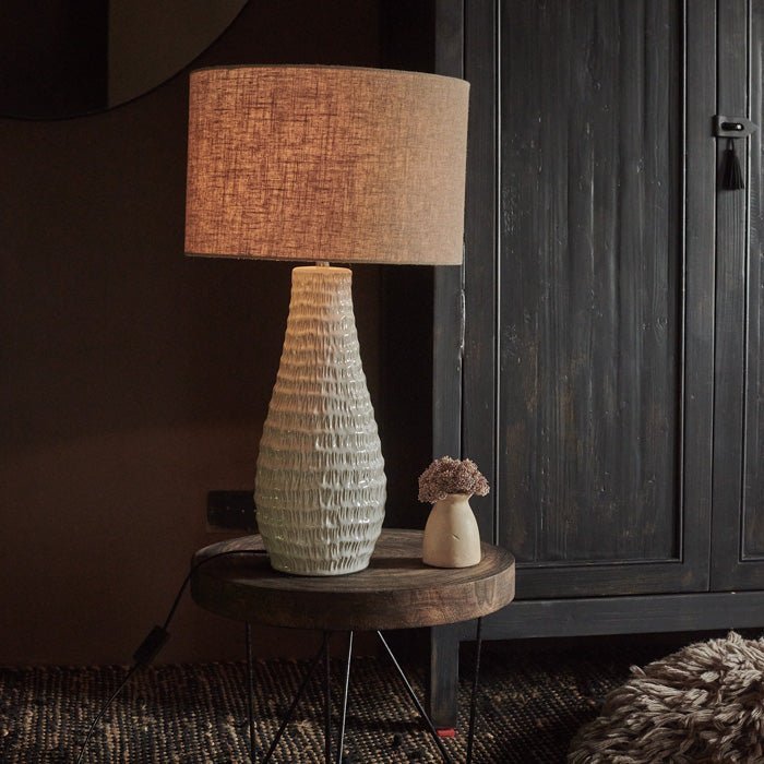 The Aspal Table Lamp, warmly lit on a low wooden side table against a warm brown wall.