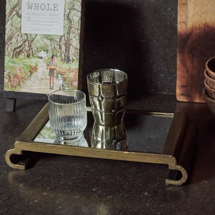 The Bletchley Mirrored Tray, a rectangular tray with a curved pedestal base sits on a black work surface next to a book.