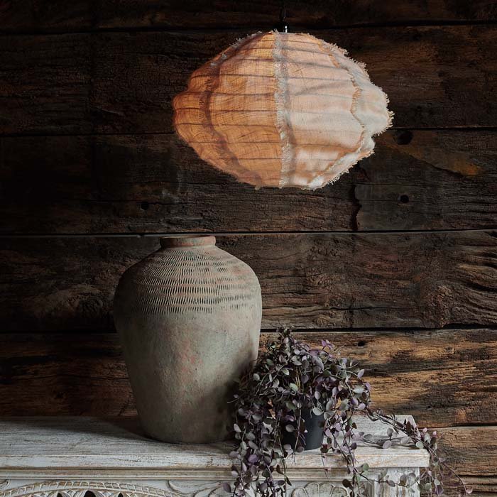 A medium sized fabric pendant lampshade hung above an ornate console with a concrete vase and an artificial houseplant.