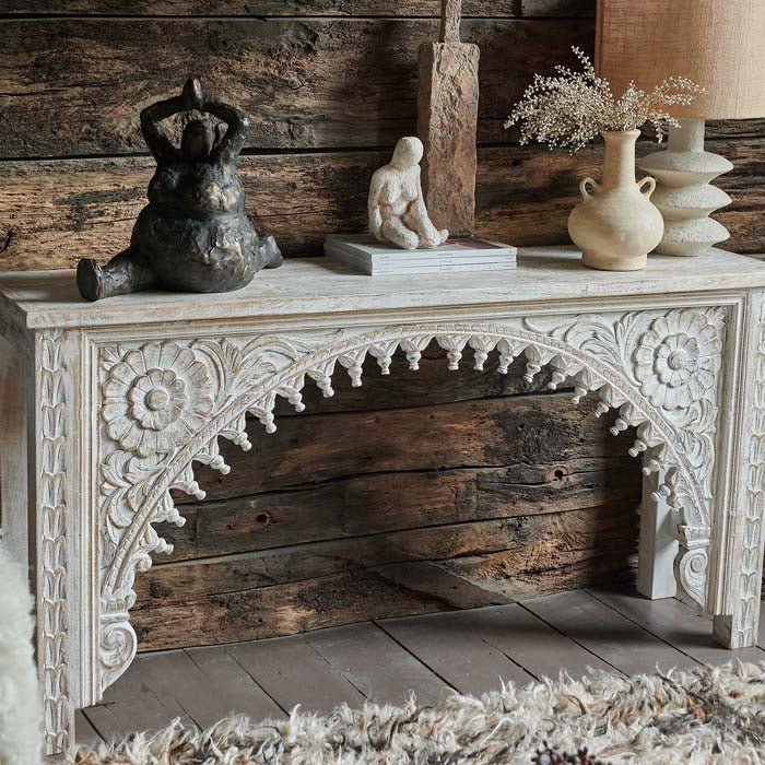 The Karnata Carved Console, a slim wooden console with an elaborately carved arched front design, with rustic accessories.