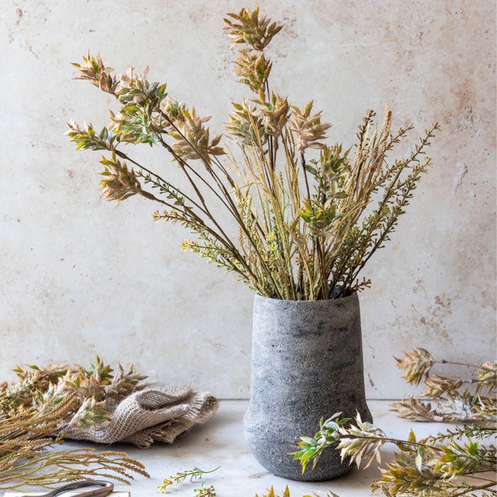Styled image of a selection of a artificial foliage arranged in a pale brown and green faux bouquet in a grey vase