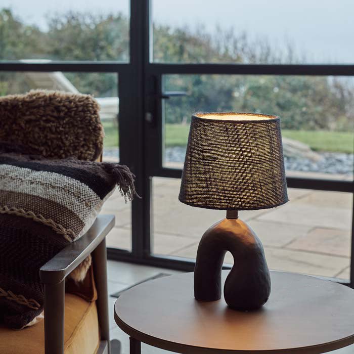 Styled image of an upside down U-shaped black ceramic lamp with a woven shade. This ceramic table lamp gives off a nice, cosy glow.
