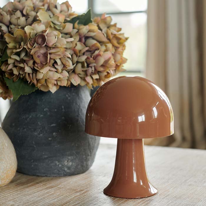 Julio LED Lamp by Abigail Ahern, known for her rule breaking designs and stylish interiors. A close up of a small portable LED table lamp in colourway called sepia which is a soft brown terracotta hue. Perfect styled on a coffee table with a vase of faux hydrangea flowers as in this vignette along with other home accessories, the perfect addition to a stylish interior. These dome shaped metal lamps are battery operated, therefore no need for plug sockets. 