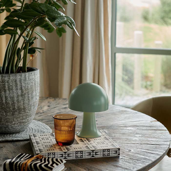 Small portable LED table lamp in forest green. Styled on a coffee table with a book and drinking glasses, it's the perfect addition to a stylish interior. These dome shaped metal lamps are battery operated, therefore no need for plug sockets. These small curvy lamps are super flexible to pop all over. the house, style next to your bath, bedside or create intrigue to your tablescapes. Available in another colourway as well as this soft timeless green colour. 