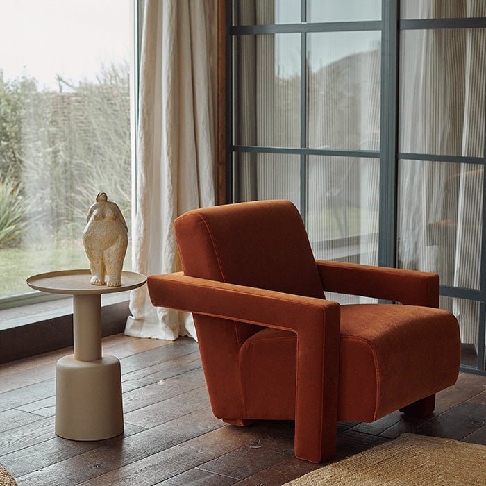 Abigail Ahern Phoenix Velvet Armchair in rich burnt orange velvet, featuring a gently reclined silhouette with angular armrests and plush upholstery, styled as a statement chair alongside a side table and decorative accessories in a warm contemporary interior.