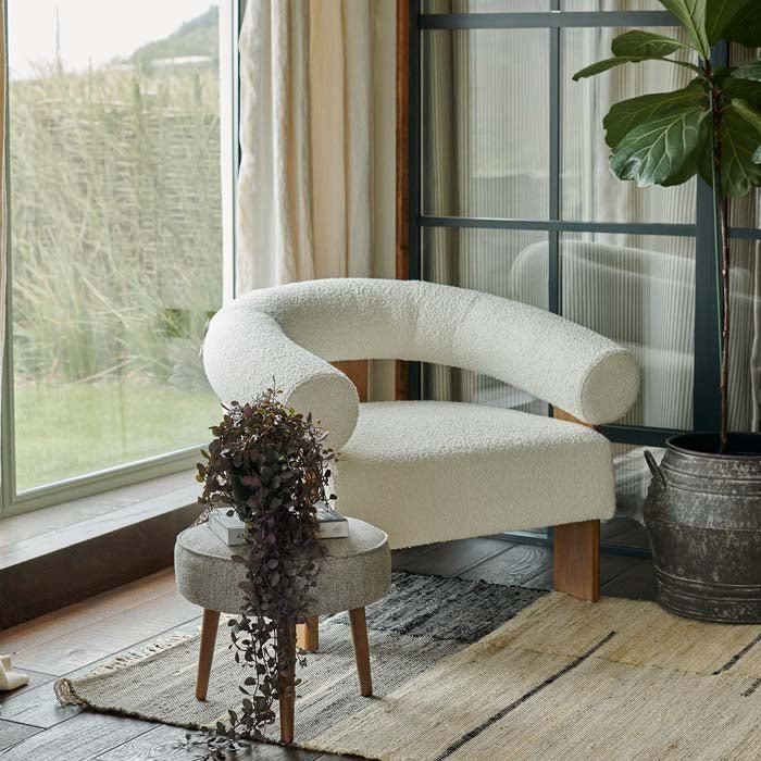 Abigail Ahern Ryton Cream Boucle Armchair with plush cream boucle upholstery, a curved backrest and distinctive cylindrical armrest, styled beside a small wooden stool and trailing plant in a warm contemporary interior.