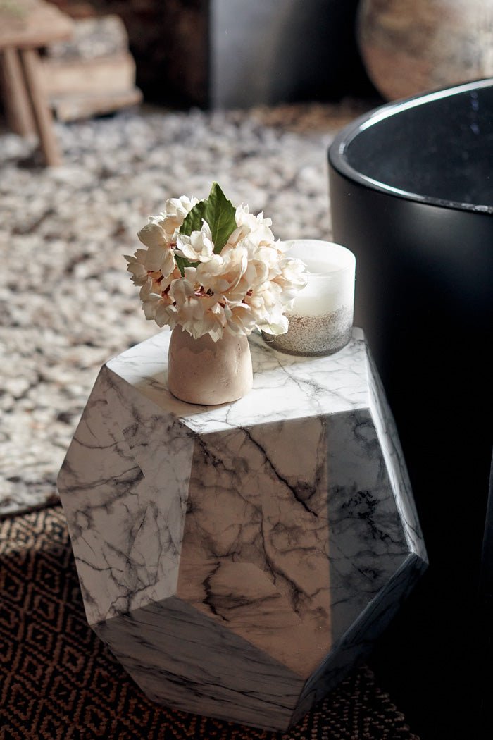 Styled image of a geometric side table with an angular shape and white marble look, styled with a faux bouquet of artificial flowers in a small vase.