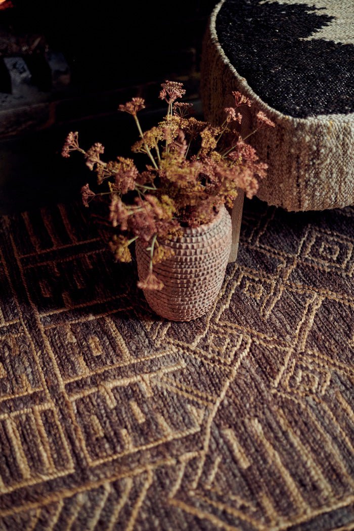 Detail image of the woven texture of a patterned brown rug with a vase of artificial flowers and an accent chair on it.