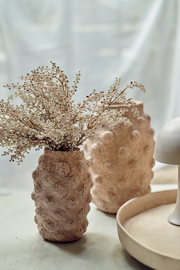 A textured beige cement vase with a bobble texture holding some artificial cream bud stems.