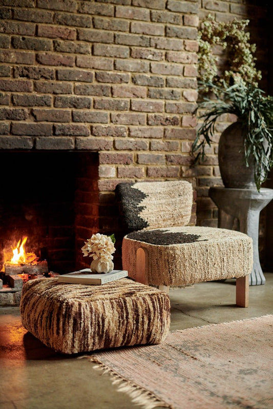 Styled image of a natural wool chair with a geometric black pattern and three wooden legs, next to a fireplace and a brown and cream striped pouffe.
