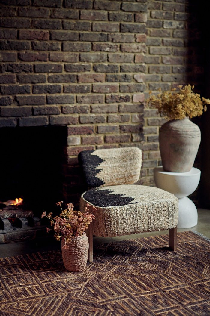 Styled image of a cream accent chair sitting on a woven brown rug next to two large vases with faux flower bouquets in front of a fireplace.