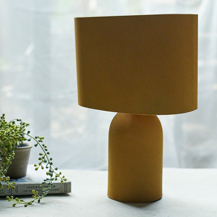 An ochre table lamp with a round base and a cylindrical shade in the same colour next to a small artificial plant.