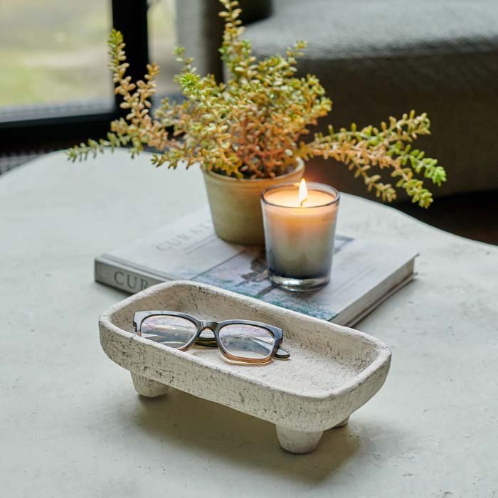 Small cream cement tray holding eye glasses on a table next to a candle and a faux plant.