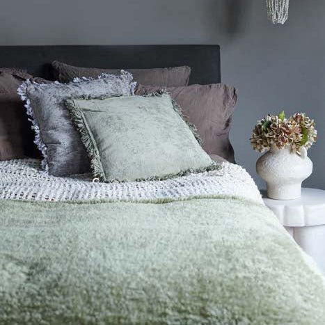 The Winona Blanket -Sage, a large super-soft and snuggly throw in a sage green hue, on a bed with some cosy cushions..