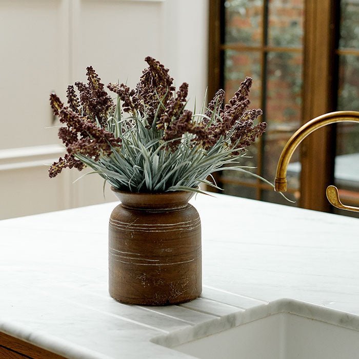 A bouquet of artificial lavender bush in a brown ceramic vase, sitting on a kitchen counter.