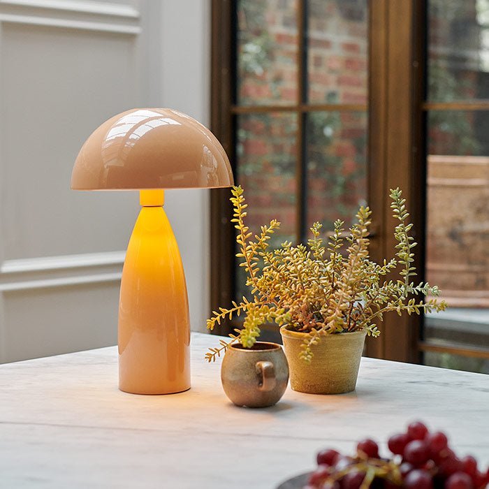 A blush pink table lamp with a dome shade and shiny finish giving off a warm glow next to a small ceramic mug and artificial plant.