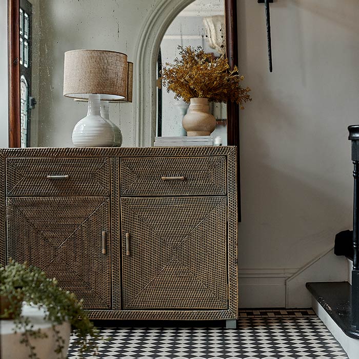 The Merida Natural Rattan Cabinet, a woven rattan cabinet with two drawers and doors, with a ceramic lamp in natural tones.