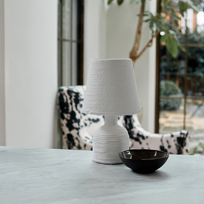 A small white table lamp made from paper mache with a ribbed design next to a bowl on a countertop.
