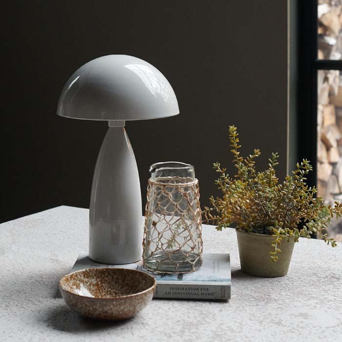 A pale grey table lamp with a dome shade and glossy finish next to some tableware and an artificial plant.