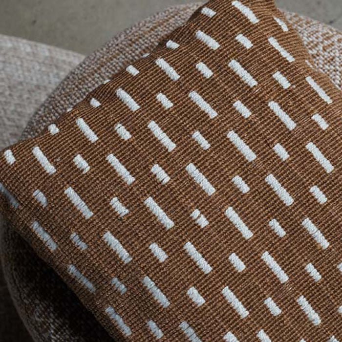A closeup of the Soriano Cushion, a square cushion with a woven cover in a rich earthy brown with white line detail.