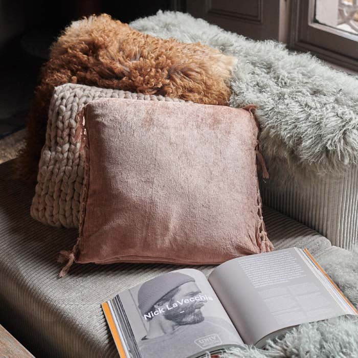The Uvero Cushion, a square fringed cushion in a dusky pink hue, next to a pale sheepskin throw and an open book on a sofa.
