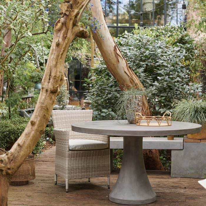 The Datura Round Dining Table, a large round cement table with a pedestal base in a leafy garden by some trees.