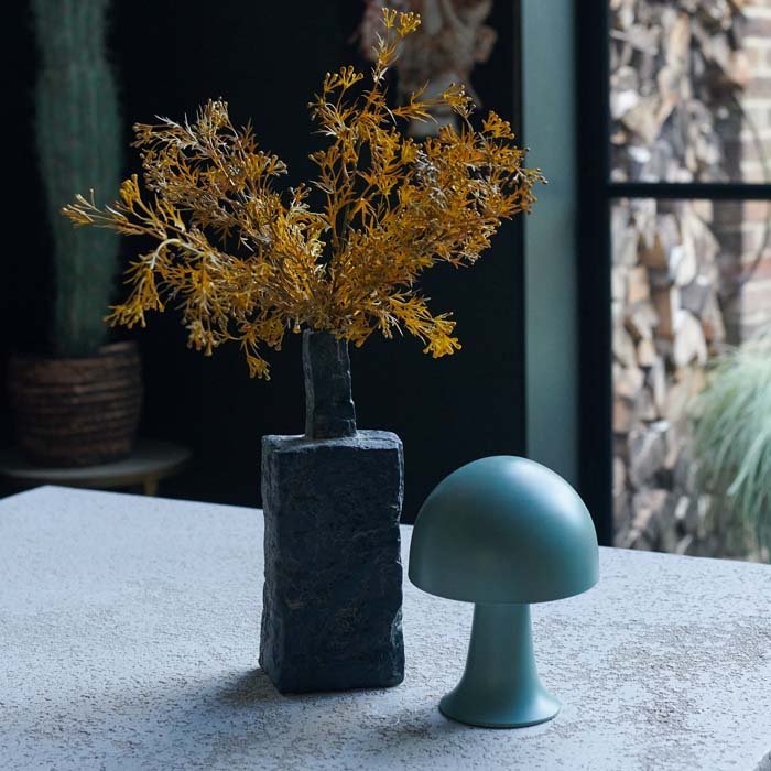 A rough textured dark grey porcelain vase in a rectangular style with some yellow artificial flowers and a small green dome table lamp.