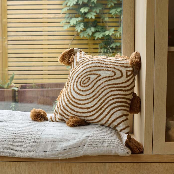 The Demelza Embroidered Cushion, a square cream coloured cushion with a radiating organic pattern and tassels in terracotta.