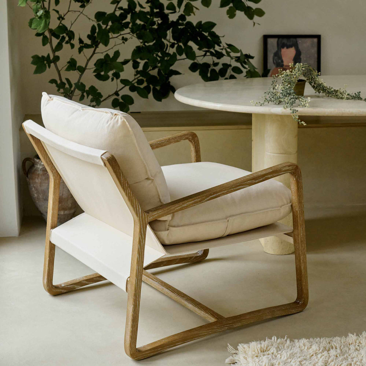 The Hockley Armchair, a modernist wooden framed armchair with cream cushioning next to a marble table and a tall plant.