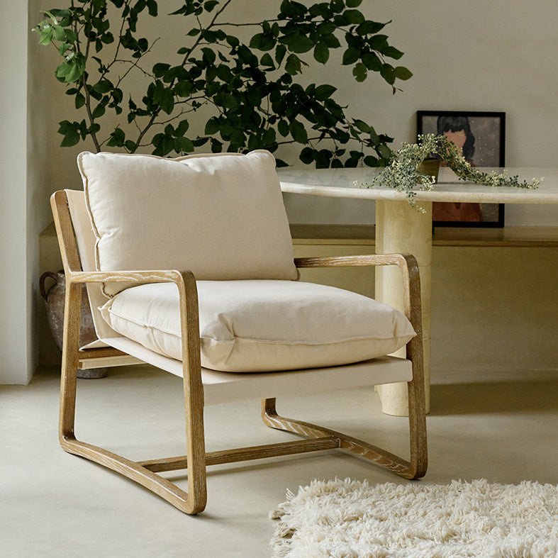The Hockley Armchair, a modernist wooden framed armchair with cream cushioning next to a marble table and a tall plant.