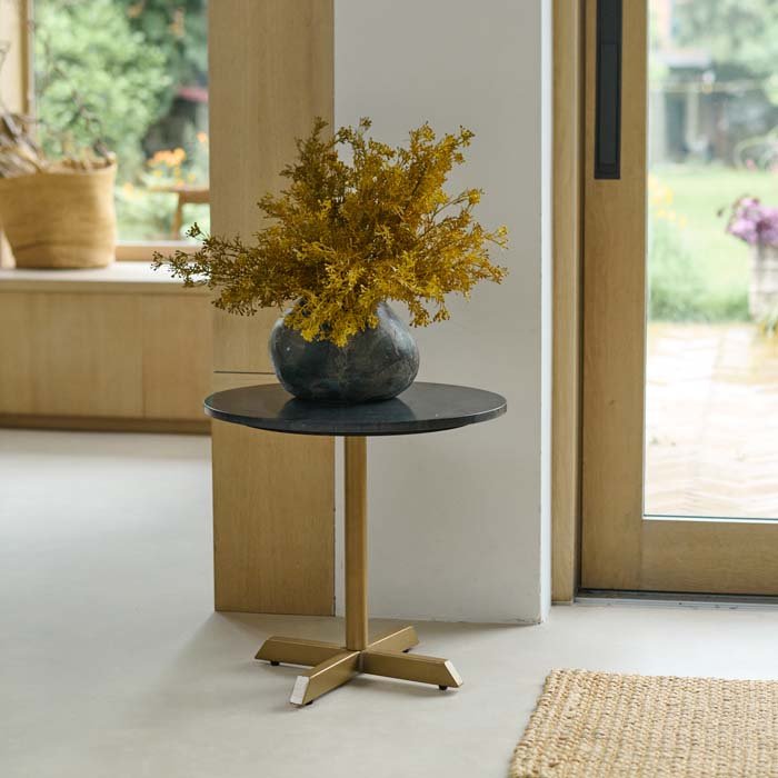 Varana Round Side Table, with a thick black marble top and a pedestal leg in gold colour with four feet, in a sunny lounge.