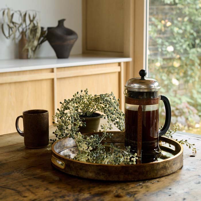 The Wilton Tray, a round tray with a mirrored surface and burnished antique gold handles, with a cafetiere and potted plant.