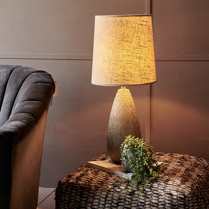 Table lamp with engraved patterned base and beige shade, displayed on woven pouffe.
