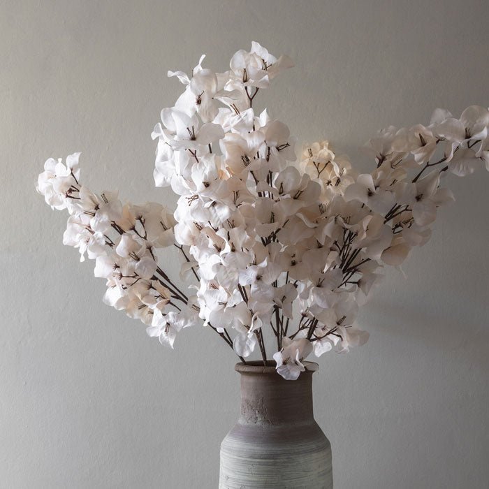 Several artificial stems of faux bougainvillea in a very pale blush colour, sitting in a grey ceramic vase.