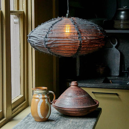 Oval shaped black fabric pendant shade switched on hanging above a wooden table with a brown jug and cooking dish. Luxury lighting to make your home look and feel good. 