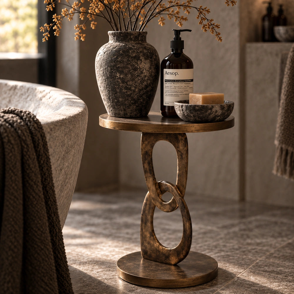 Emilio Side Table styled beside a bed with a textured vase, amber bottles and small objects, showing its sculptural linked base in a neutral bedroom.