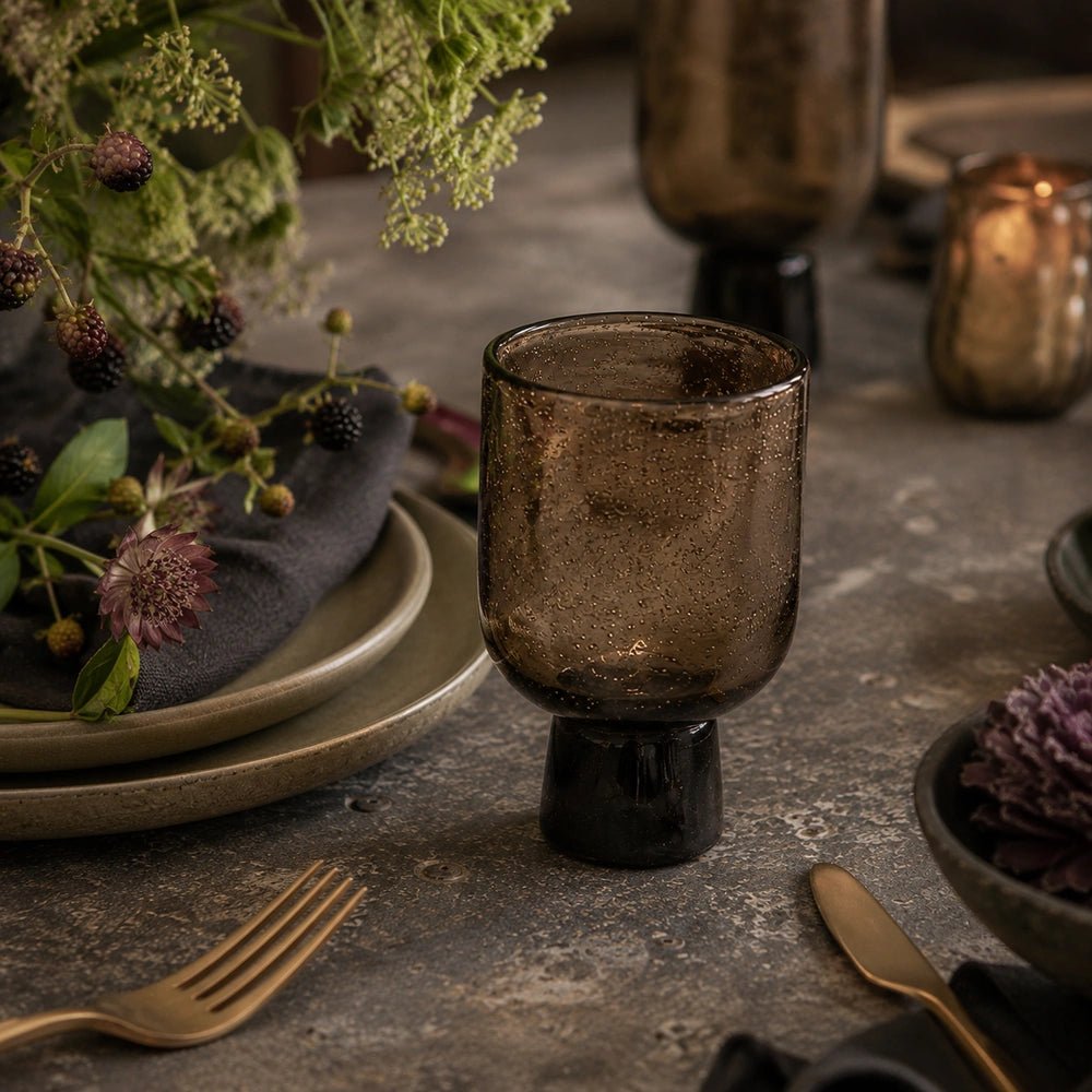 Evie Glass, Burnt Umber with warm brown textured bowl and dark pedestal base, styled among plates, flowers and candlelight on a dining table.