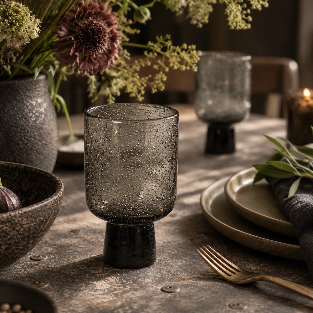 Evie Glass, Smoke shown close up in a dining table setting, with its textured smoky bowl and dark pedestal surrounded by patterned tableware and flowers.