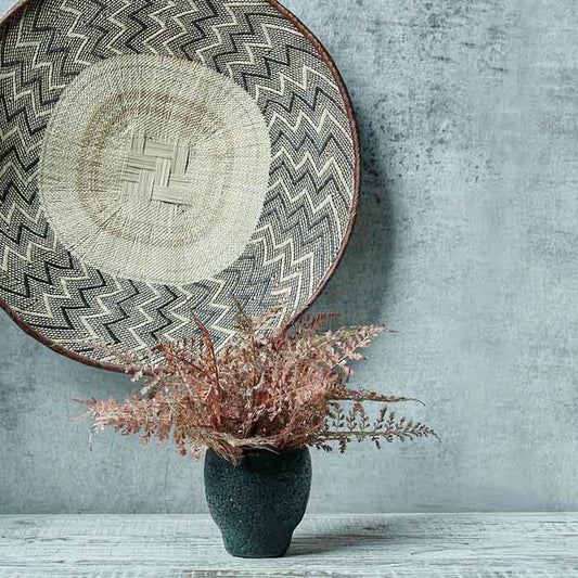 Large round woven basket with a zig zag pattern hung on a grey wall above orange fern stems in a black vase