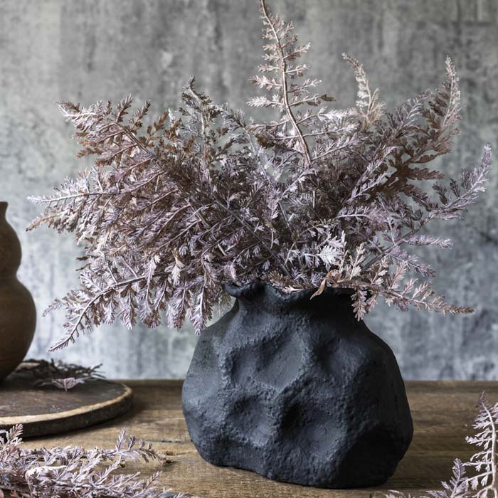A black ceramic vase holding a faux bouquet of stems of artificial purple fern..
